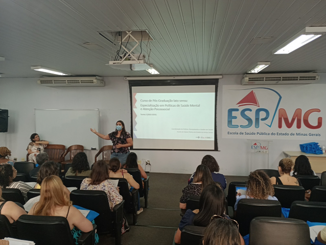 Imagem mostra uma sala de aula onde está acontecendo o curso. Alunos estão de costas sentados nas carteira e na frente a uma professora e uma projeção no quadro.